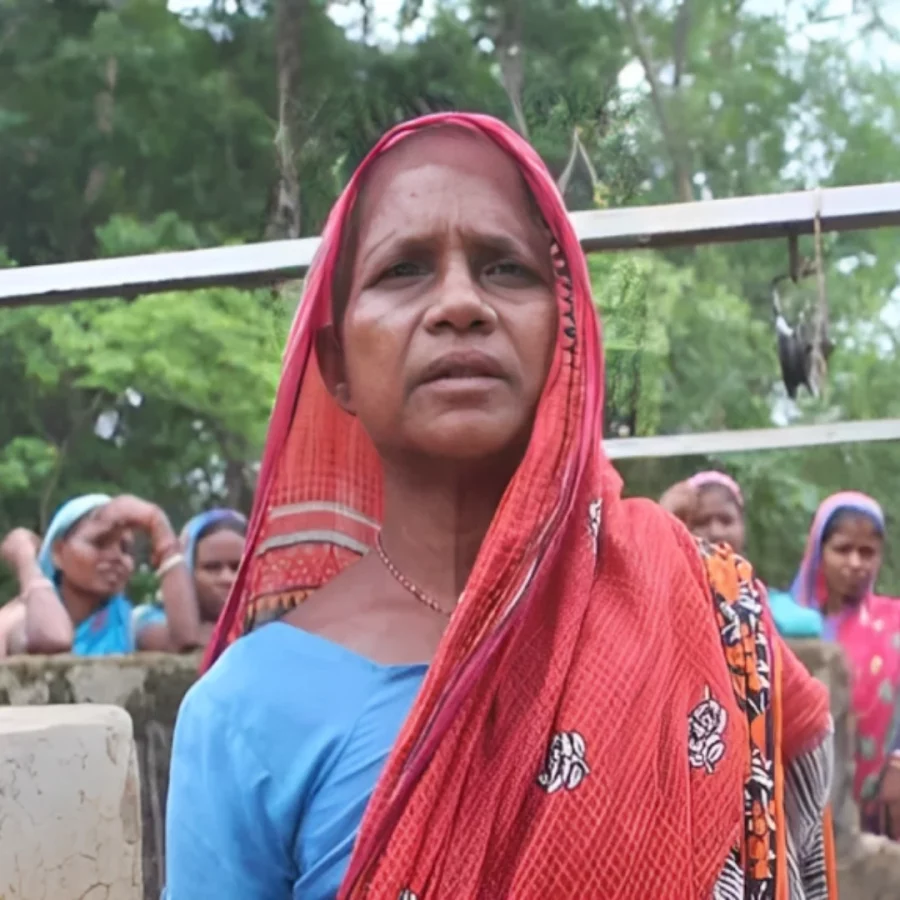 A village woman in red saree looking at the camera with a few other women in the background.